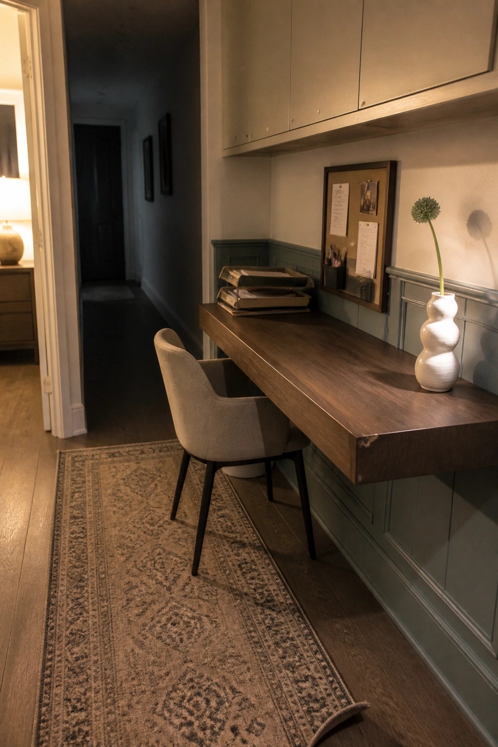 A long wooden wall-mounted desk sits below light-colored upper cabinets in a narrow interior space, with a gray upholstered chair on a patterned rug and a corkboard on the wall.