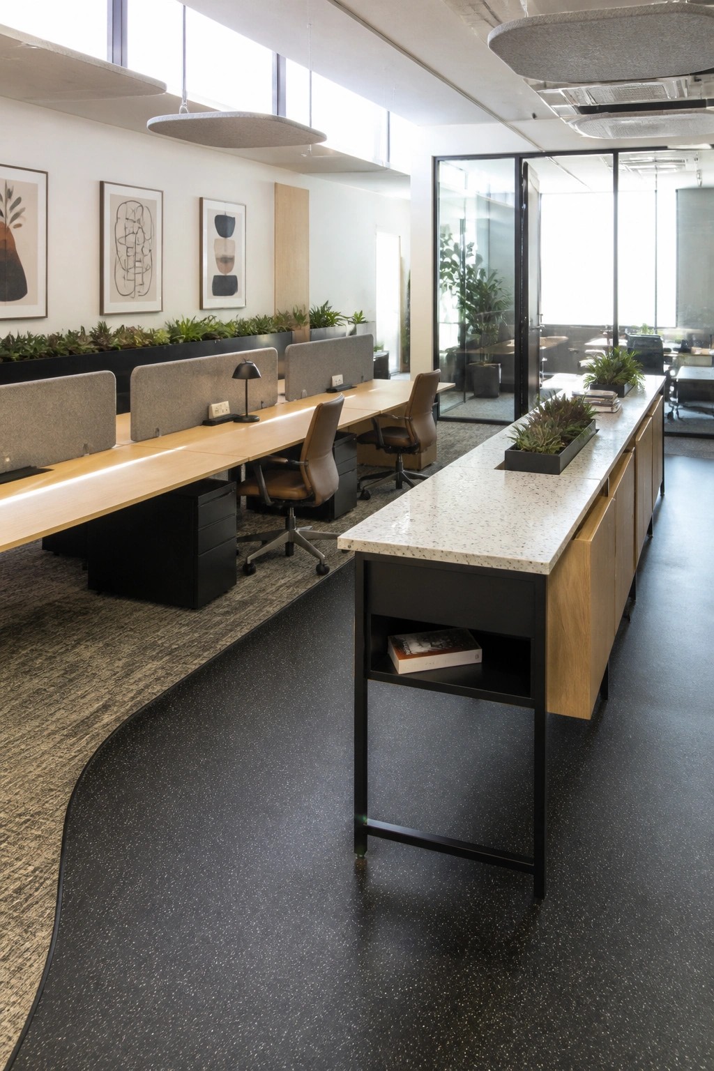 An open office interior with long wooden desks along the wall divided by gray partitions and a central island table with black legs and wood cabinets.