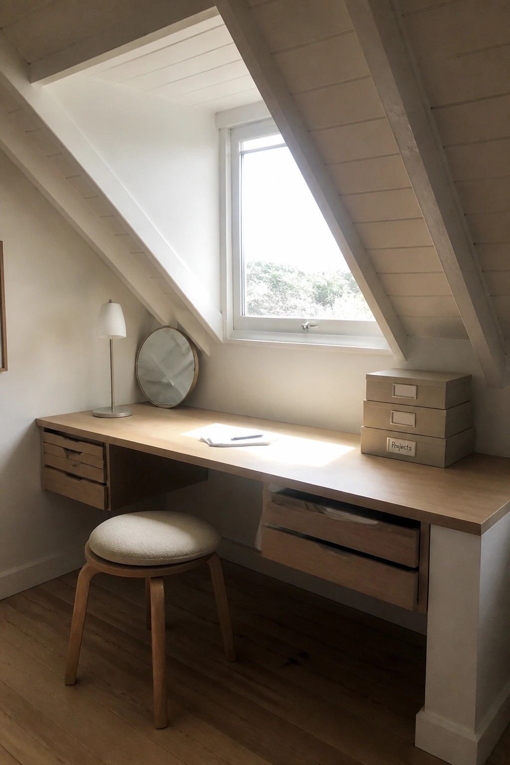 A light wood desk with built-in drawers sits beneath a slanted ceiling next to a window, with a round stool in front and only a few small items on the surface.