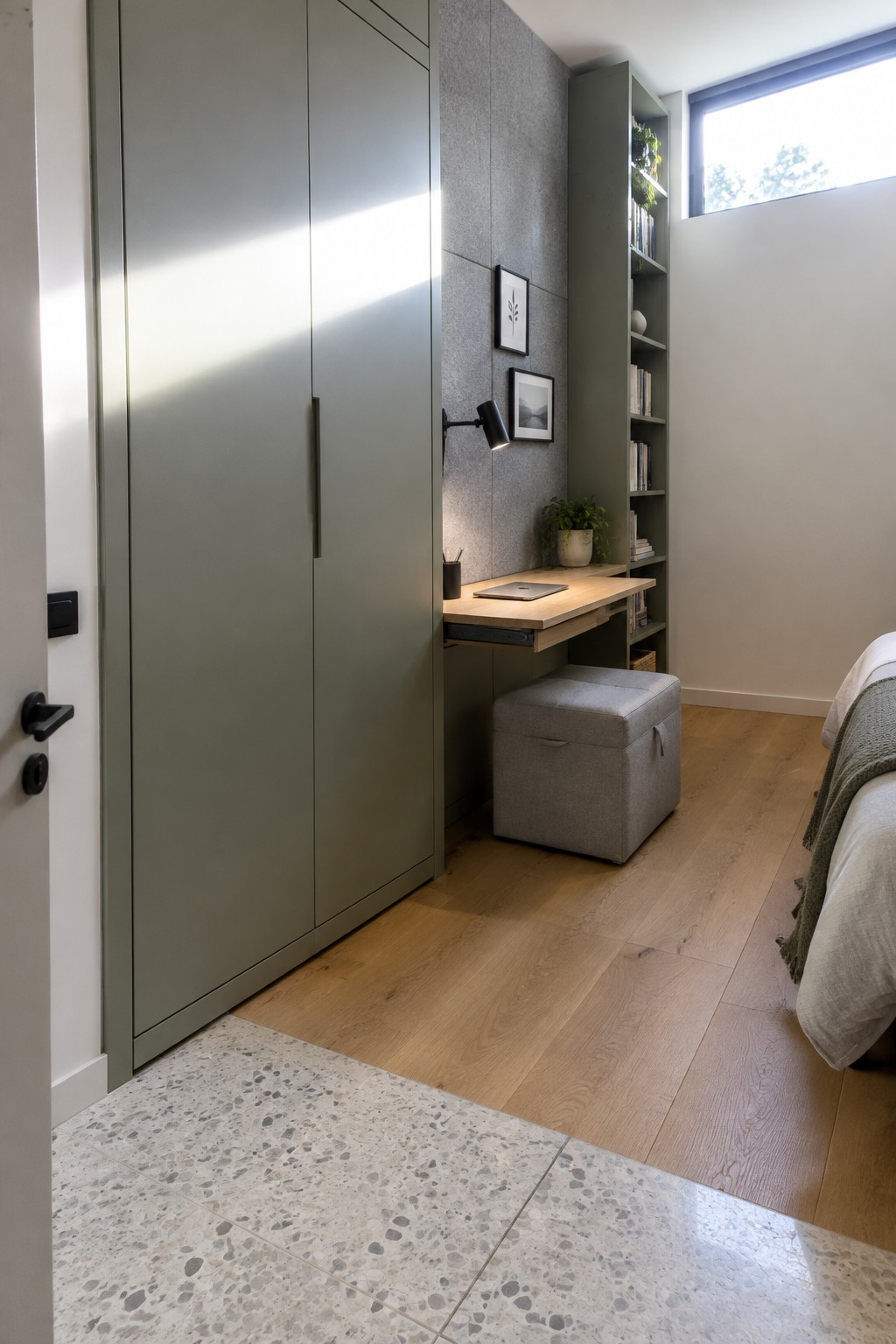 A bedroom interior showing a tall green wardrobe beside a wooden fold-out desk with a laptop on it, a gray storage ottoman underneath, and built-in shelving on the adjacent wall.