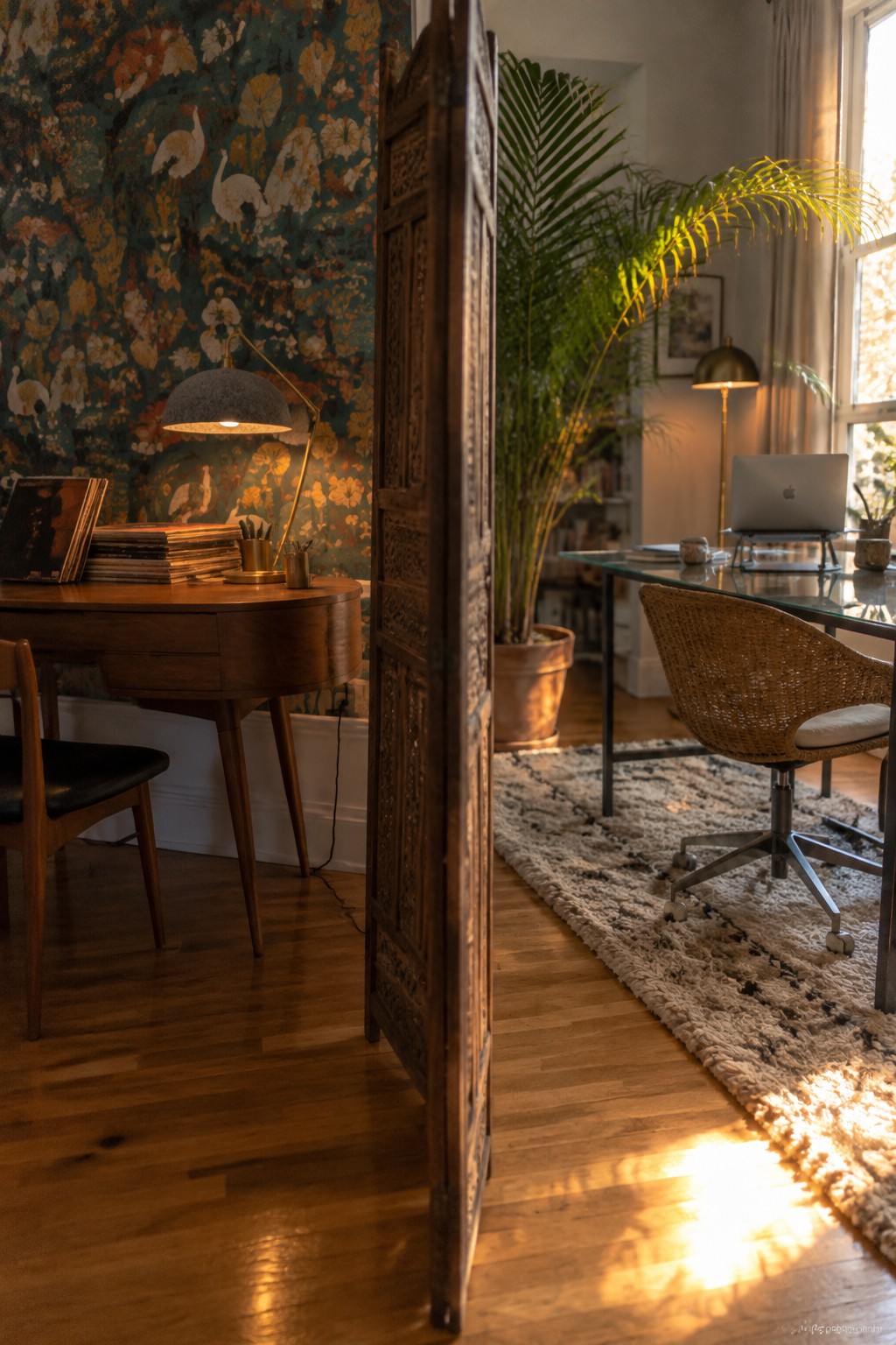 A home office divided by a tall carved wooden screen, with one wooden desk against floral wallpaper on the left and a glass desk with a laptop on the right.
