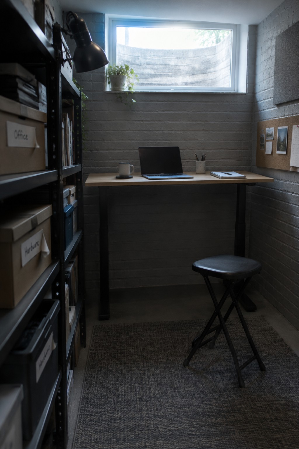A narrow home office with a wooden desk placed under a window, holding a laptop and mug, a black stool in front, and metal shelving with boxes on the left.