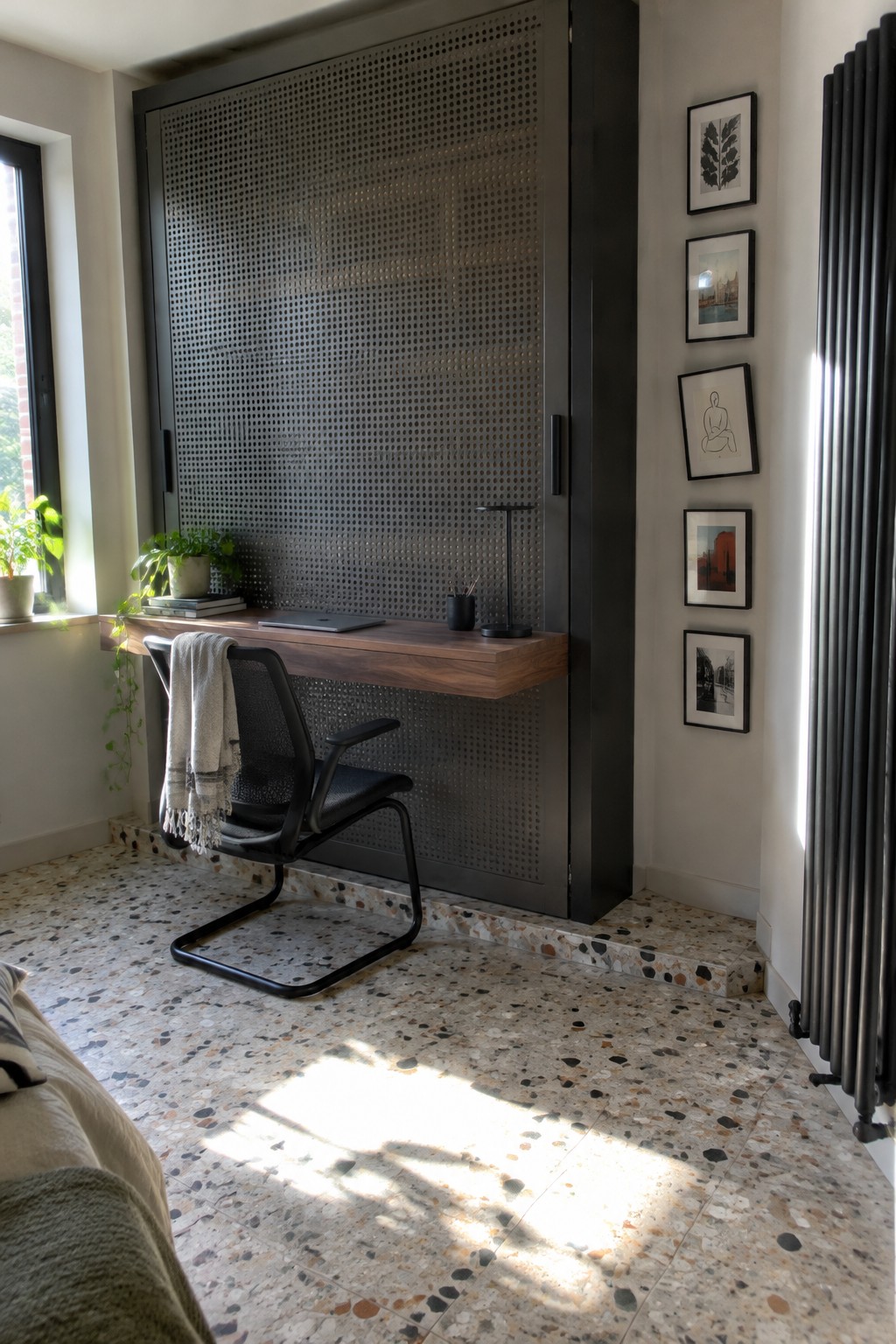 A bedroom interior showing a narrow floating wooden desk mounted in front of a tall black perforated metal cabinet, with a black mesh office chair and terrazzo floor visible.