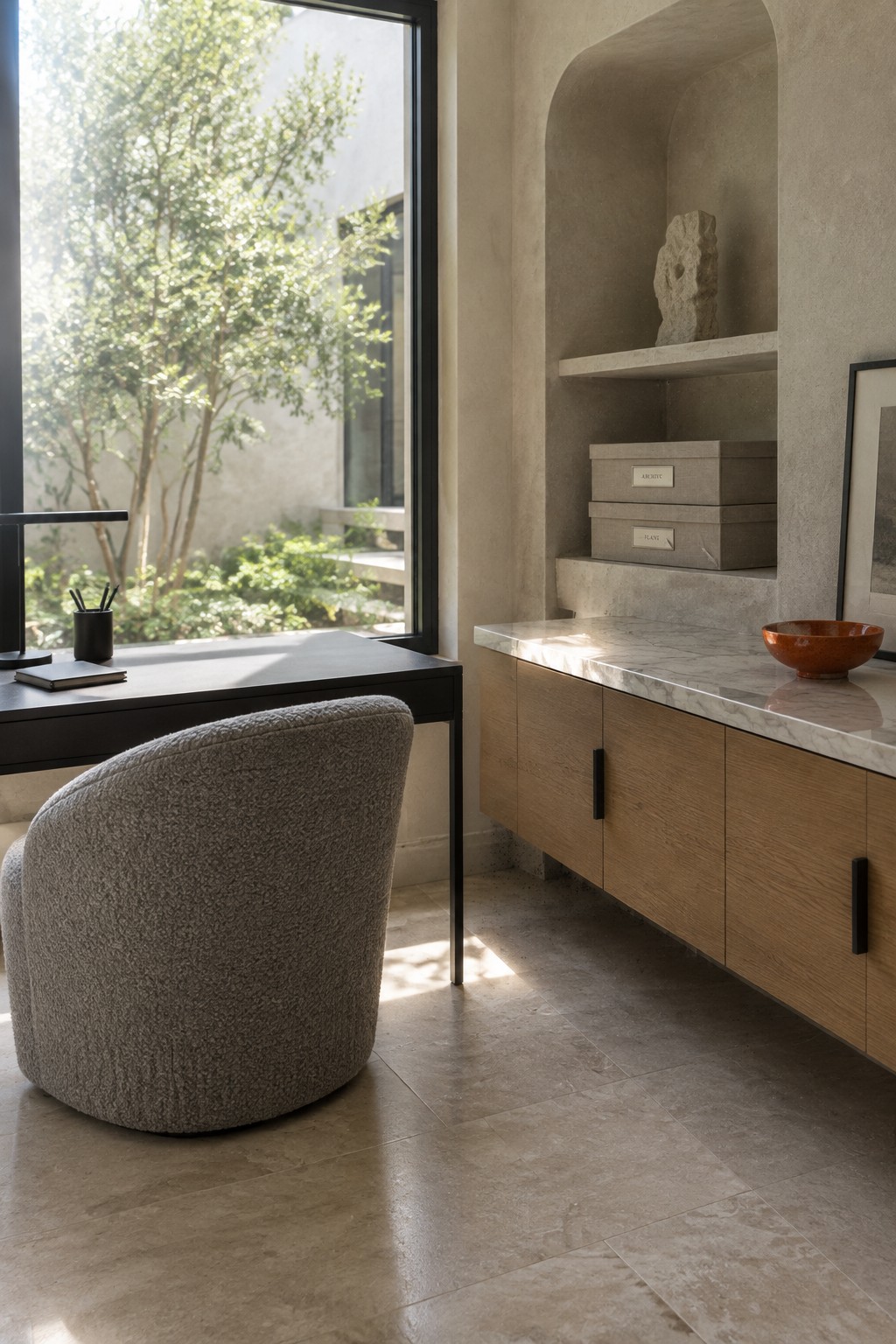 A modern home office with a black desk and gray upholstered chair facing a large window, beside a light wood cabinet topped with marble and featuring built-in open shelving above.