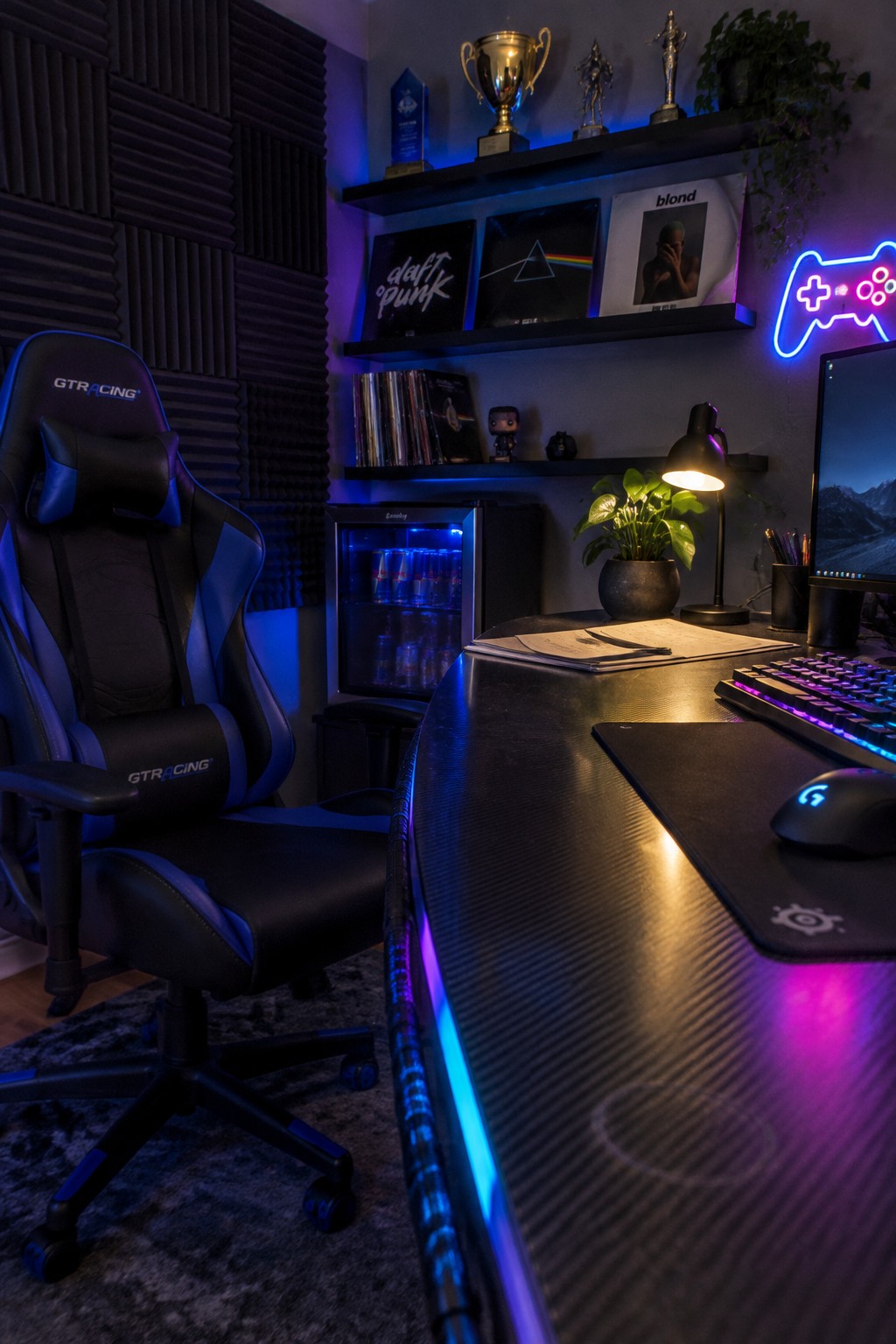 A dark home office featuring a black desk with blue and purple LED lighting along the edge, a black and blue gaming chair, and shelves holding vinyl records and trophies.