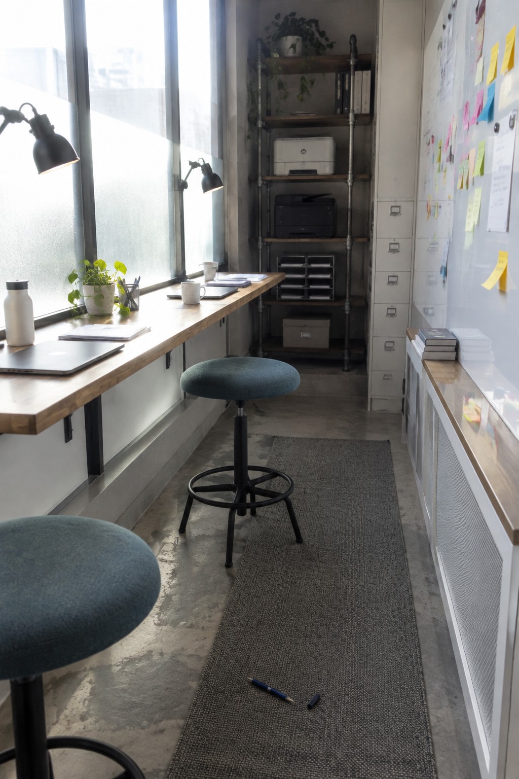 A narrow home office with a long wooden desk under large windows, two teal stools, industrial pipe shelving with a printer, and a whiteboard covered in notes and sticky notes.