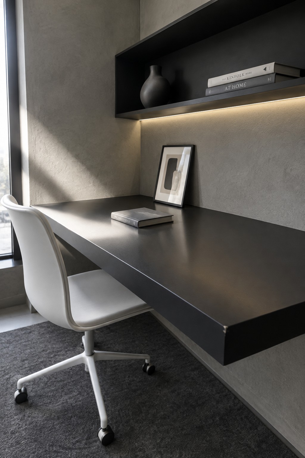 Minimalist black floating desk with white chair under illuminated shelf with books and vase
