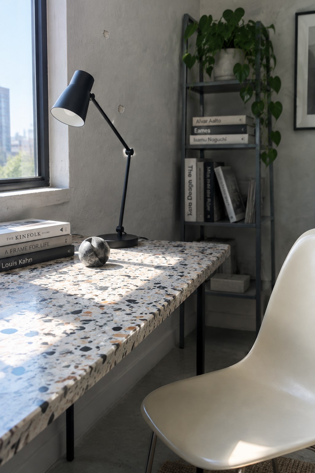 A compact home office corner with a terrazzo desk positioned under a window, featuring a black adjustable lamp, stacked books, a marble object, and a white molded chair next to a metal shelving unit with plants and books.