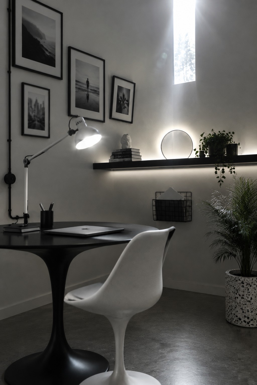 A modern home office featuring a round black desk with a white chair, an adjustable white task lamp, a laptop, and a floating shelf with backlighting holding stacked books, a stone sculpture, and potted plants.
