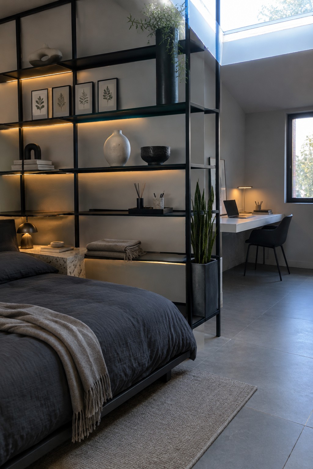 A bedroom divided by a tall black open shelving unit, with a bed on one side and a white desk with chair on the other.