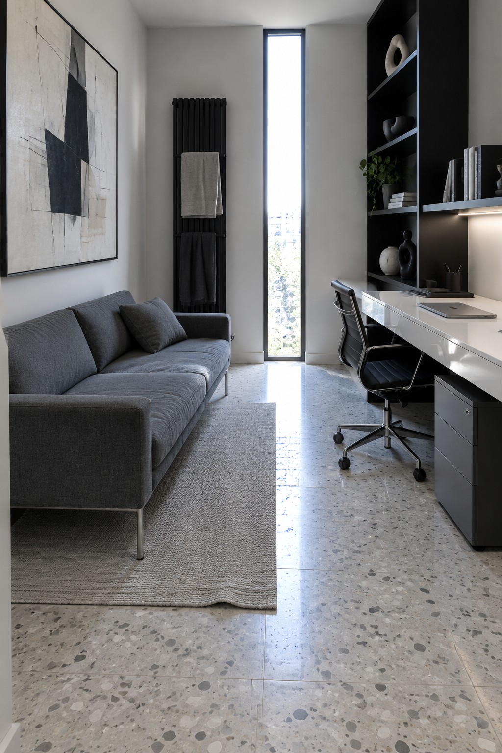 A modern home office with a gray fabric couch on a light rug, a white desk with black chair, black shelving, and a tall narrow window.