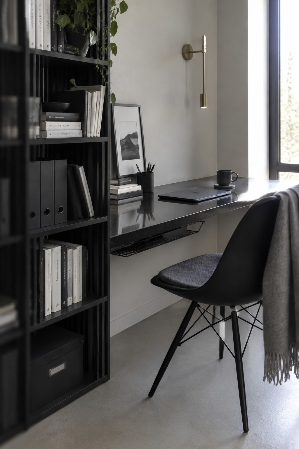 A minimalist home office featuring a floating black desk with a laptop, a black chair with gray cushion, and a tall black bookshelf filled with books and storage boxes.
