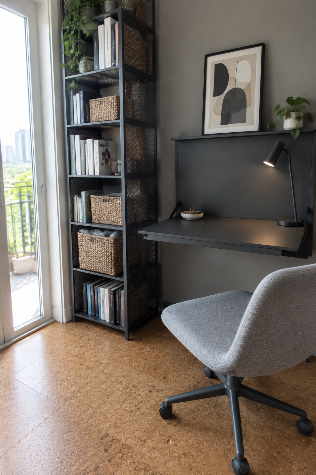 A minimalist home office corner with a dark wall-mounted fold-down desk, tall black mesh shelving unit filled with books and baskets, gray upholstered office chair, black desk lamp, and abstract framed art on the wall.
