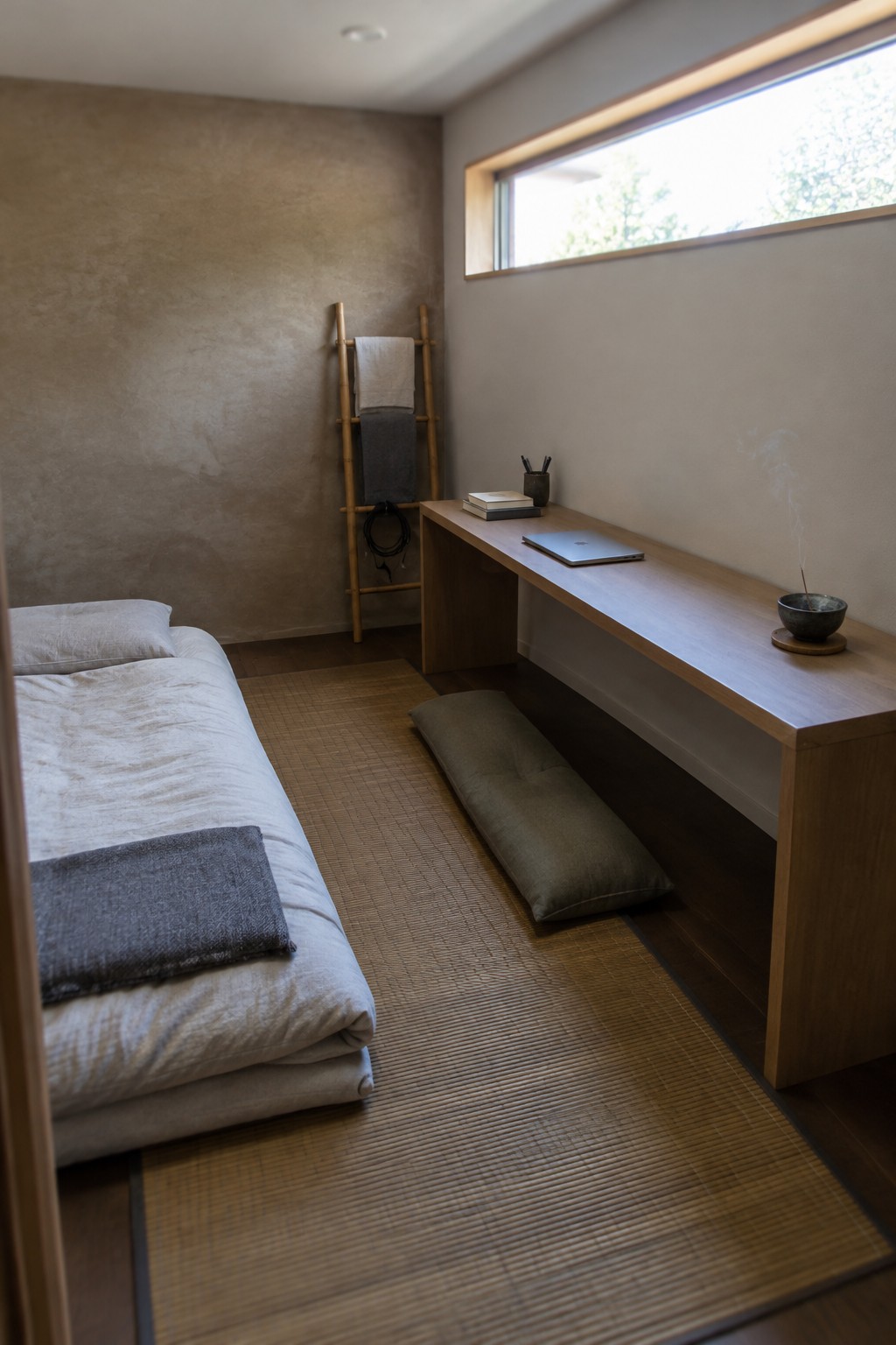 A narrow minimalist bedroom with a low bed on tatami mats on the left and a long wooden desk running along the right wall under a window, with a gray floor cushion in front of the desk.
