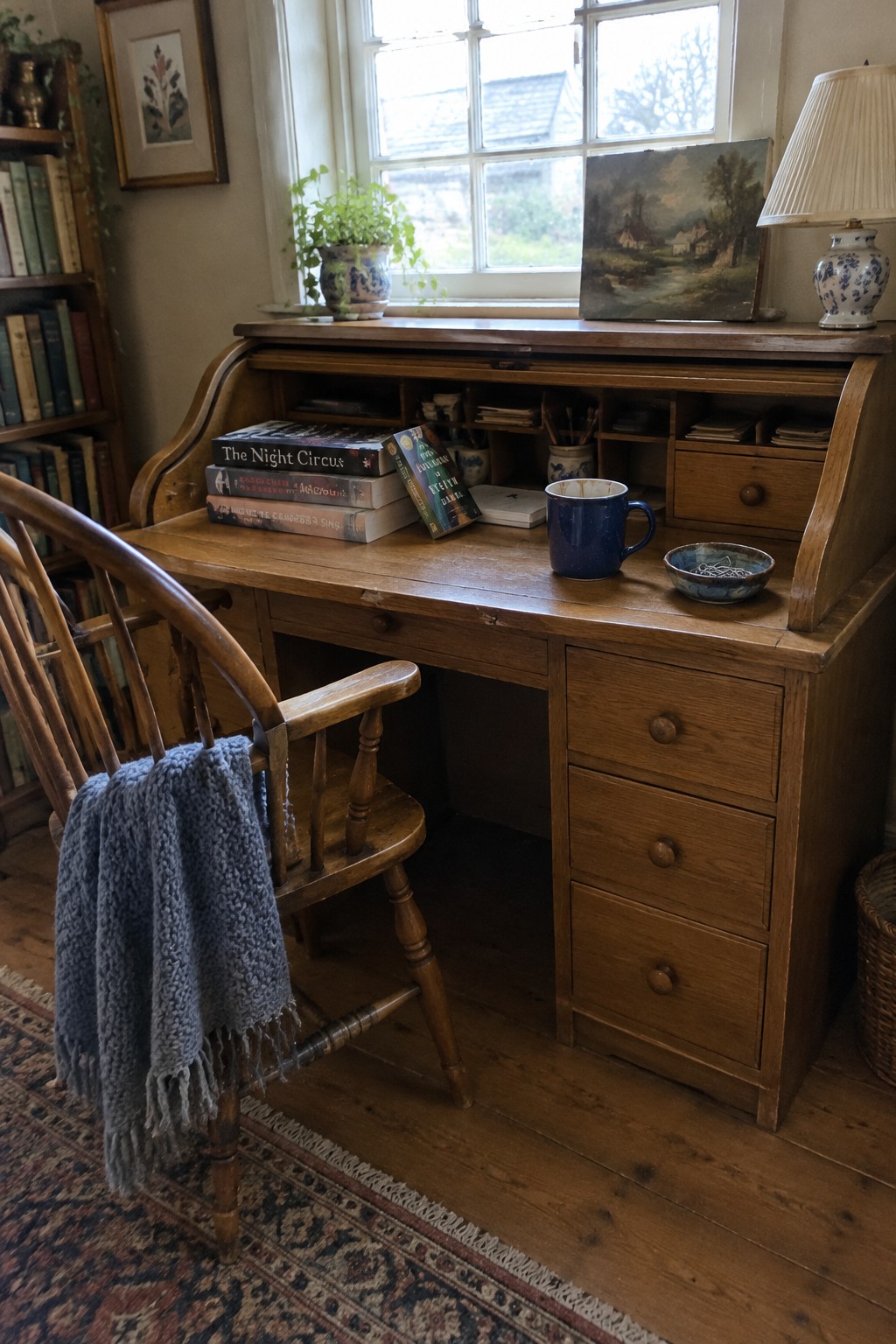 A wooden roll top desk with open compartments and drawers stands in front of a window, with a wooden chair draped in a gray blanket beside it.