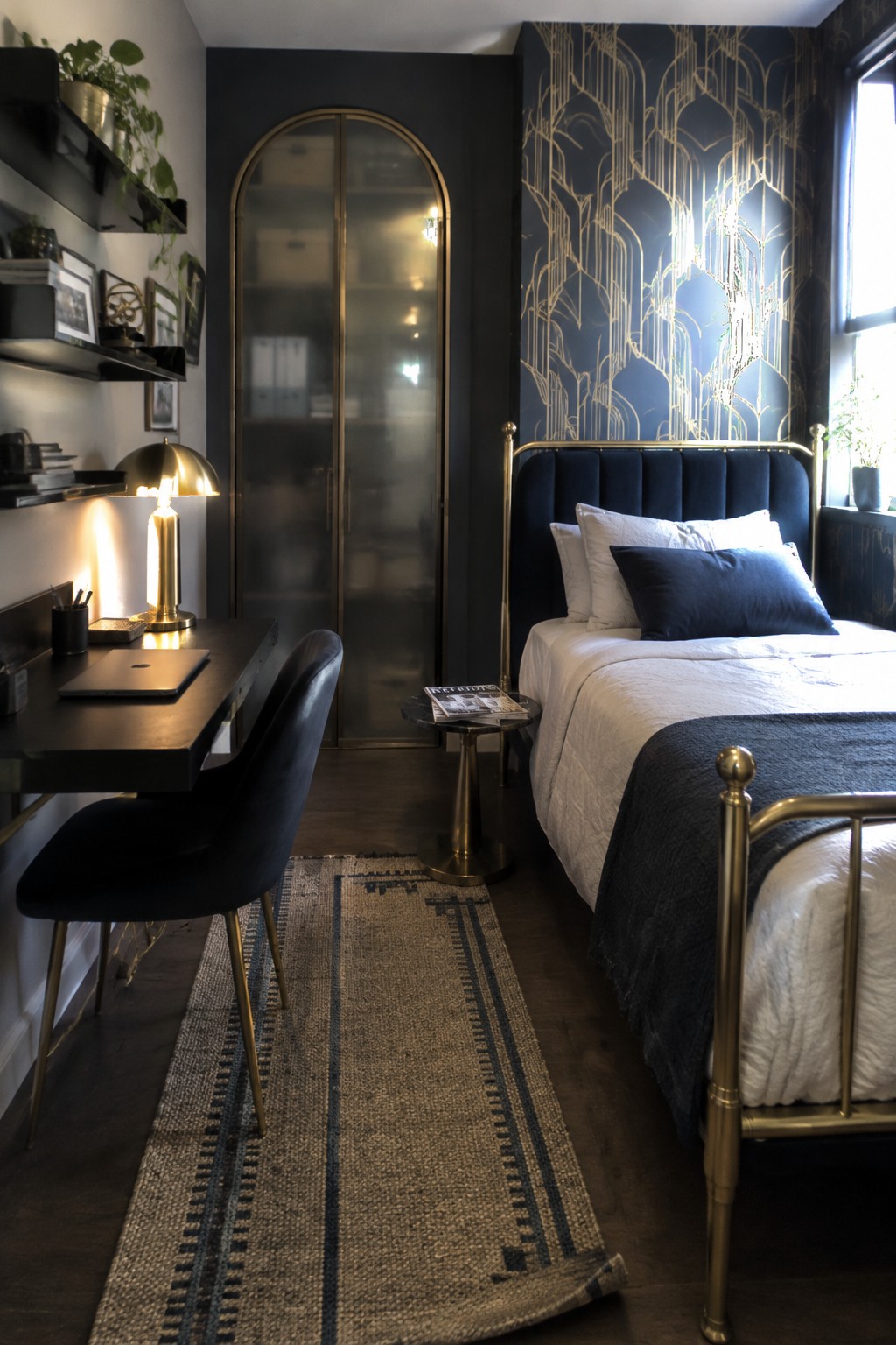 A narrow bedroom with a brass bed on the right side and a dark desk with a velvet chair on the left, separated by a long striped runner rug on dark wood flooring.