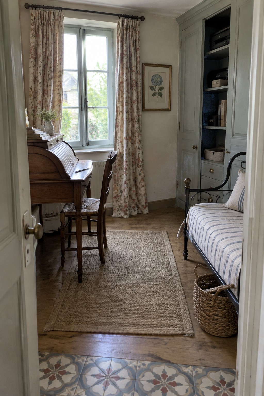 A bedroom interior showing a wooden roll top desk by a window with floral curtains, a woven rug on the floor, a striped daybed, and a tall light green cabinet.