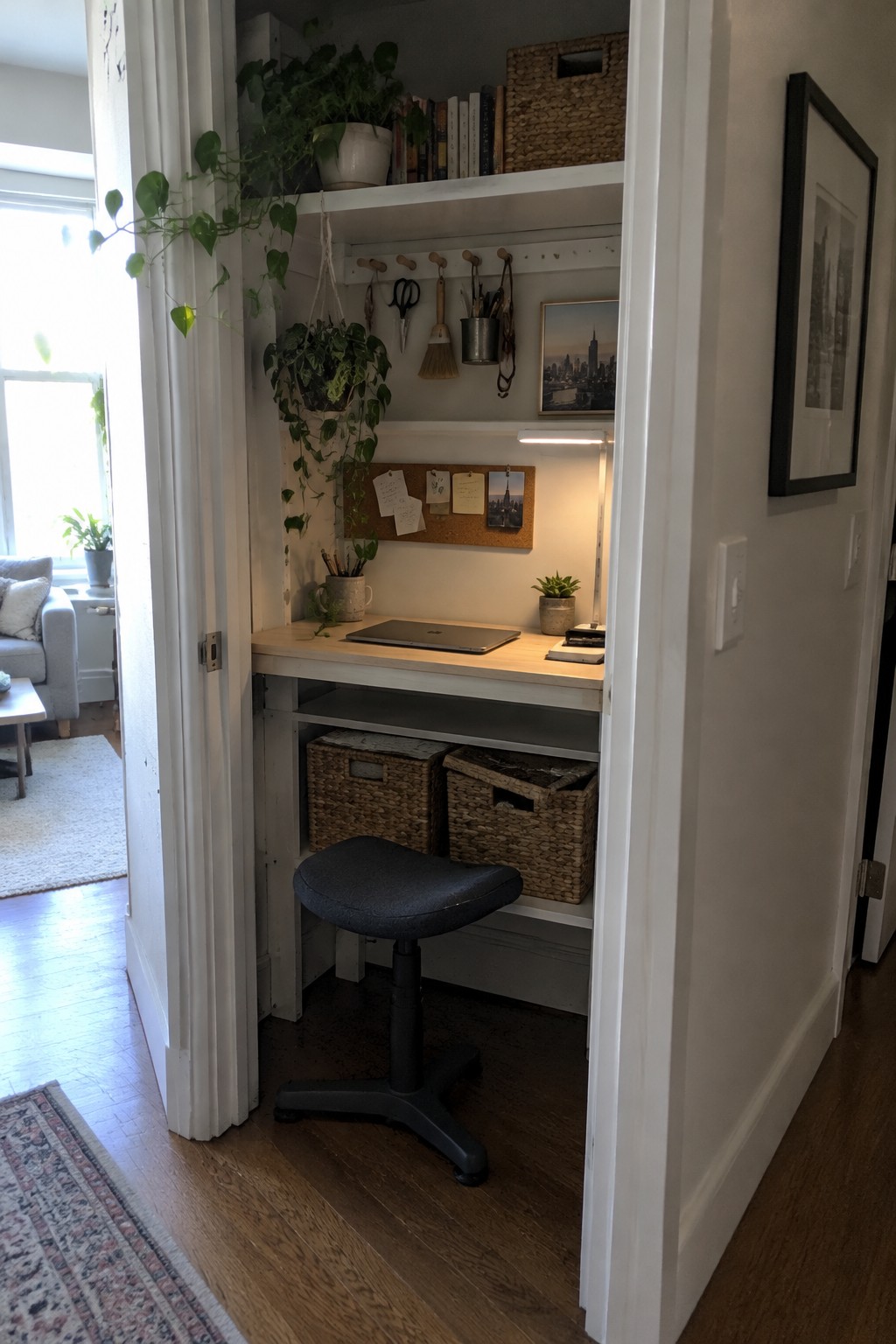 A small built-in desk inside a white closet alcove with a laptop, stool, hanging plants, books on shelves, and woven storage baskets below.