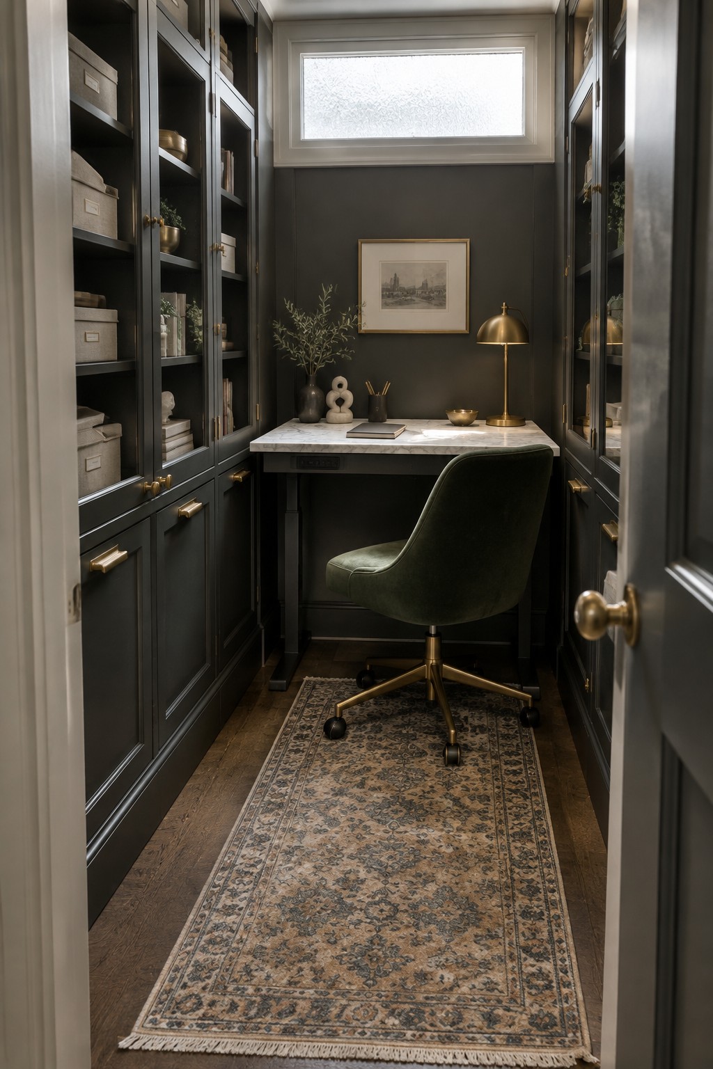 A narrow interior room with dark built-in cabinets lining both walls, a marble desk centered beneath a small window, a green upholstered office chair on a patterned rug, and brass hardware details.