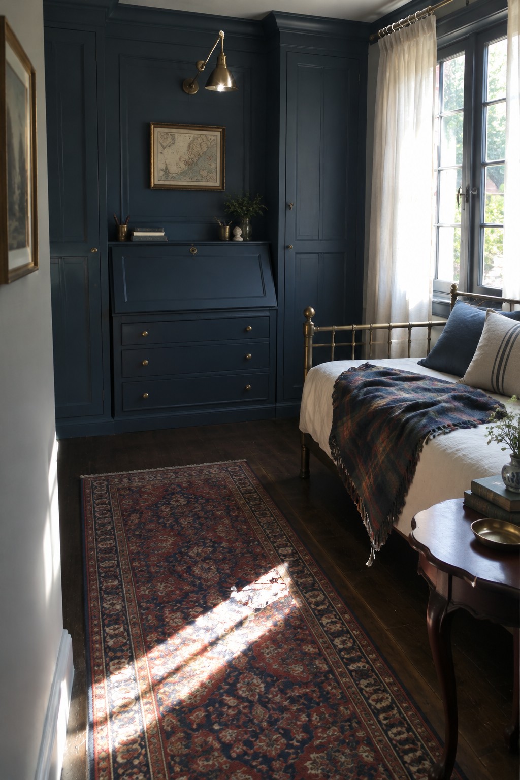 A bedroom with dark blue built-in cabinetry featuring a closed drop-down desk section, a brass bed with white bedding and a plaid throw, and a red patterned rug on dark wood floors.