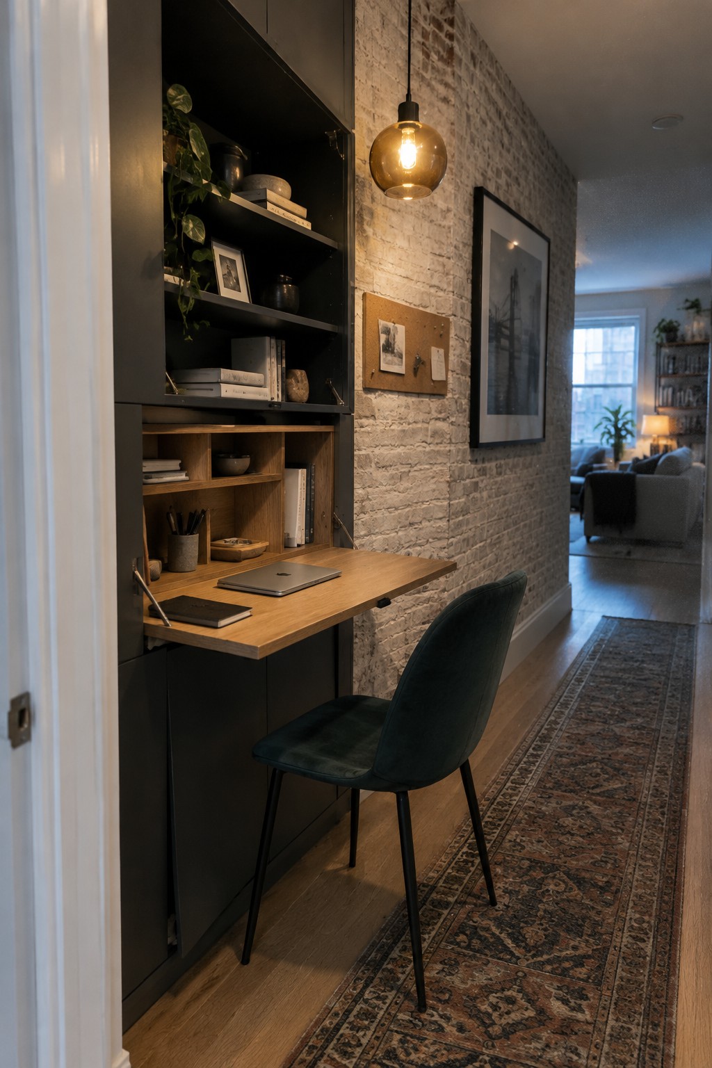 A narrow home office nook with a wooden pull-out desk extended from a dark built-in cabinet, a laptop and notebook on the desk, and a green upholstered chair in front of an exposed brick wall.