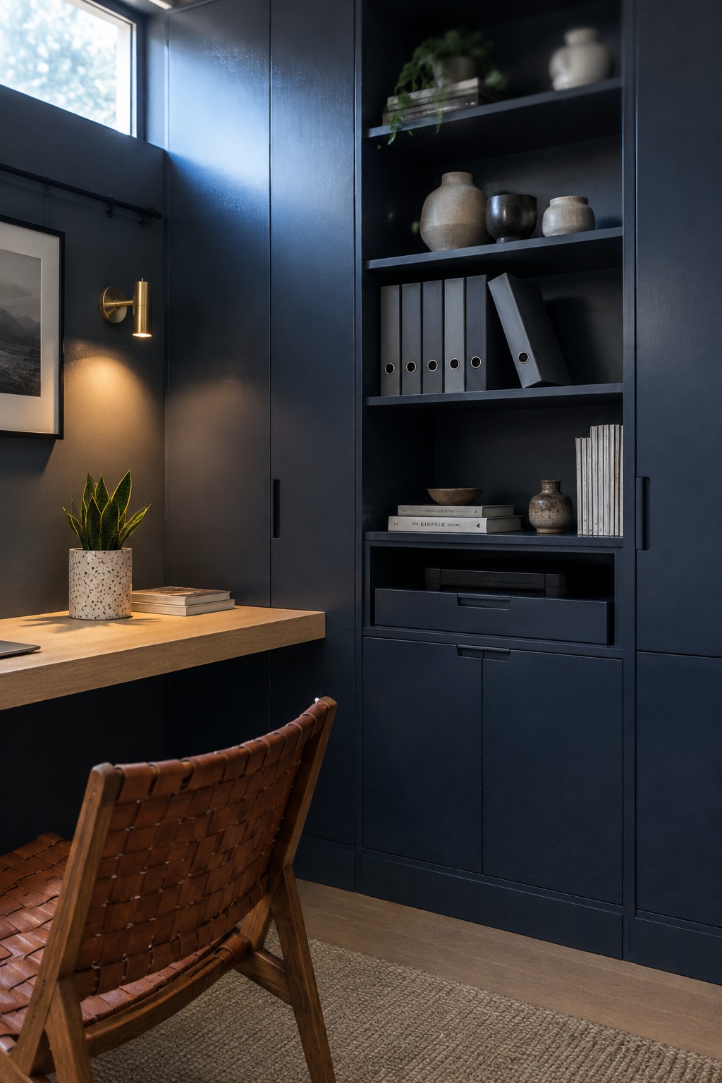 A small home office features a dark navy built-in storage unit with open shelves, drawers, and cabinets next to a light wood floating desk and woven leather chair.