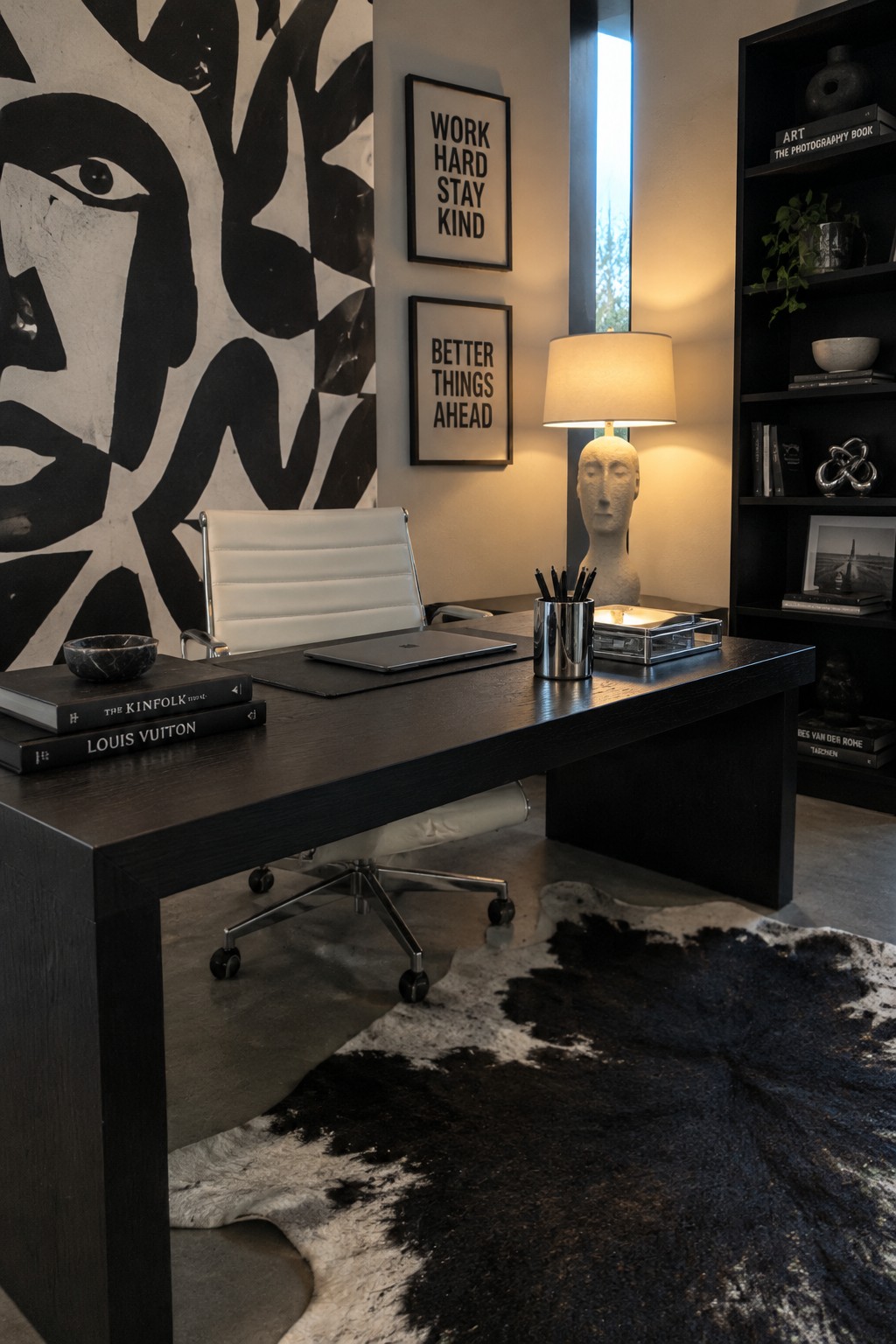A home office featuring a large black and white abstract face wallpaper on the main wall, a dark wood desk with a white leather chair, and framed text prints above the desk.