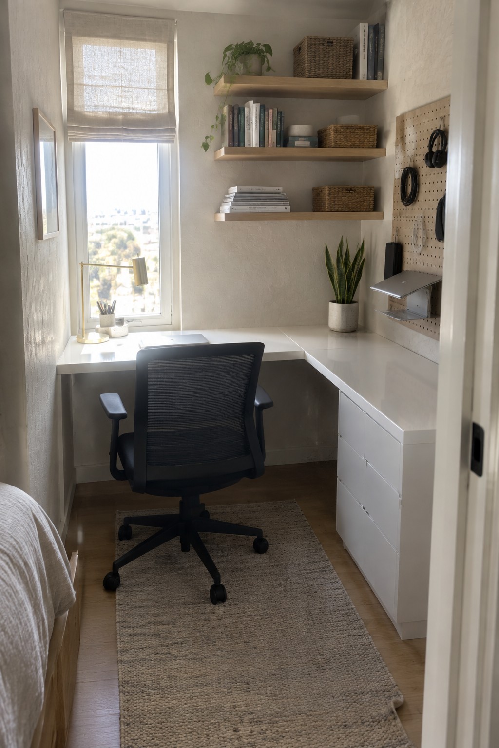 A compact home office with a white L-shaped desk in a corner under a window, a black mesh office chair on a woven rug, floating wooden shelves with books and baskets, and a pegboard mounted on the right wall.