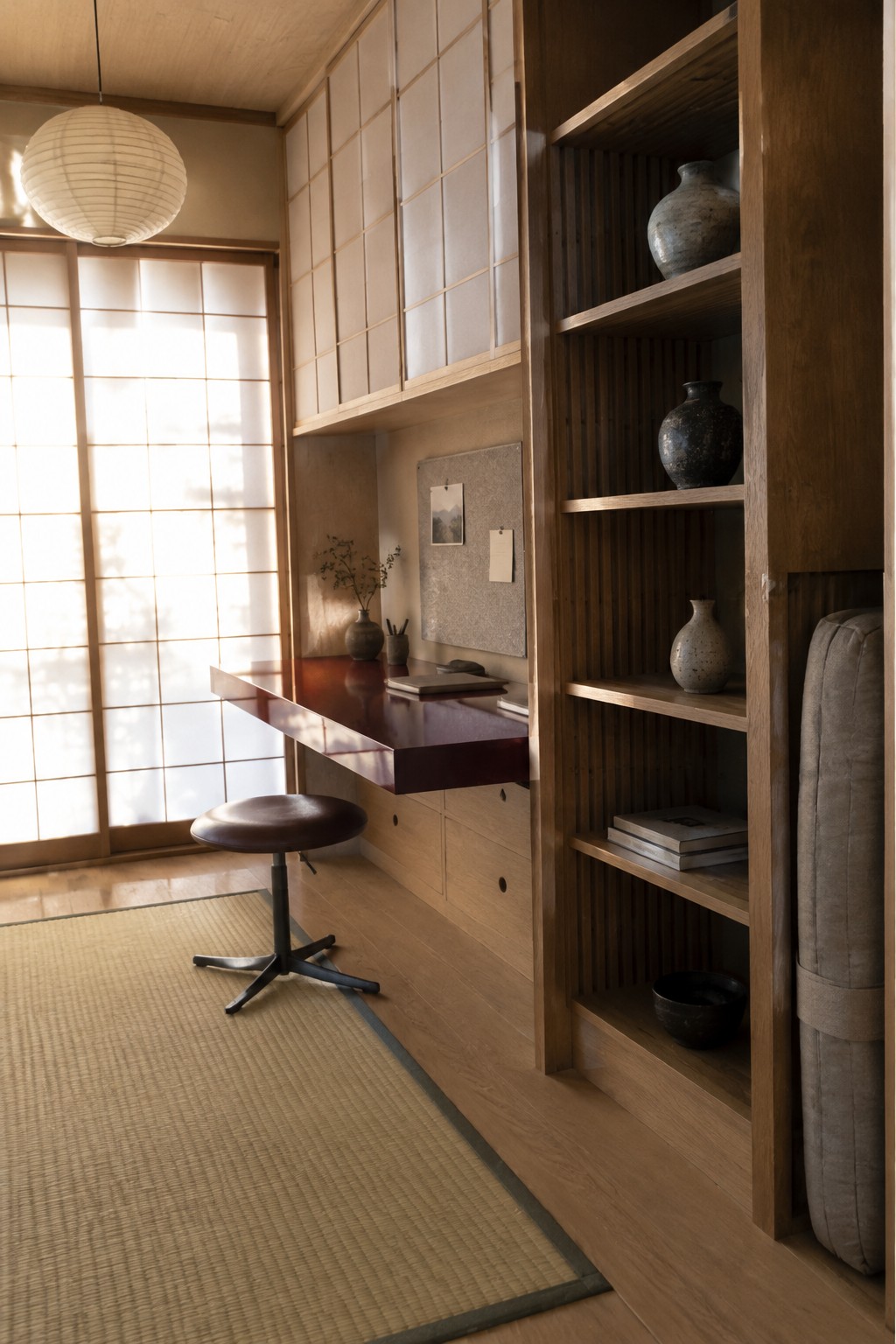A compact Japanese-style room with a wall-mounted wooden desk, swivel stool, and tall built-in shelving unit holding ceramic vases and stacked books beside shoji screens.