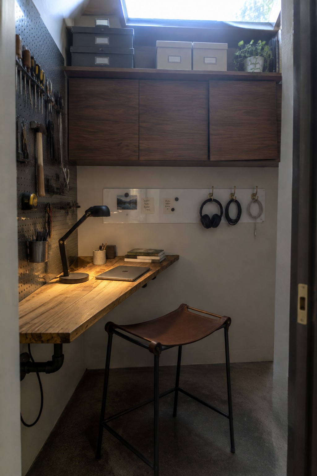A narrow home office features a wooden floating desk with a laptop and lamp, a leather stool underneath, dark wood upper cabinets, and a pegboard with tools on the left wall.