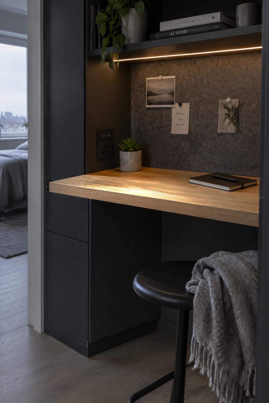 A wooden desk built into a dark closet alcove with a fabric pinboard on the back wall, under-shelf lighting, a small potted plant, and a black stool.