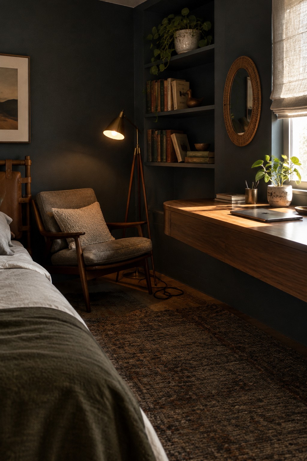A bedroom corner featuring a floating wooden desk beneath a window, an upholstered armchair with a pillow, a tripod floor lamp, built-in shelves with books and plants, and a woven mirror on a dark blue wall.