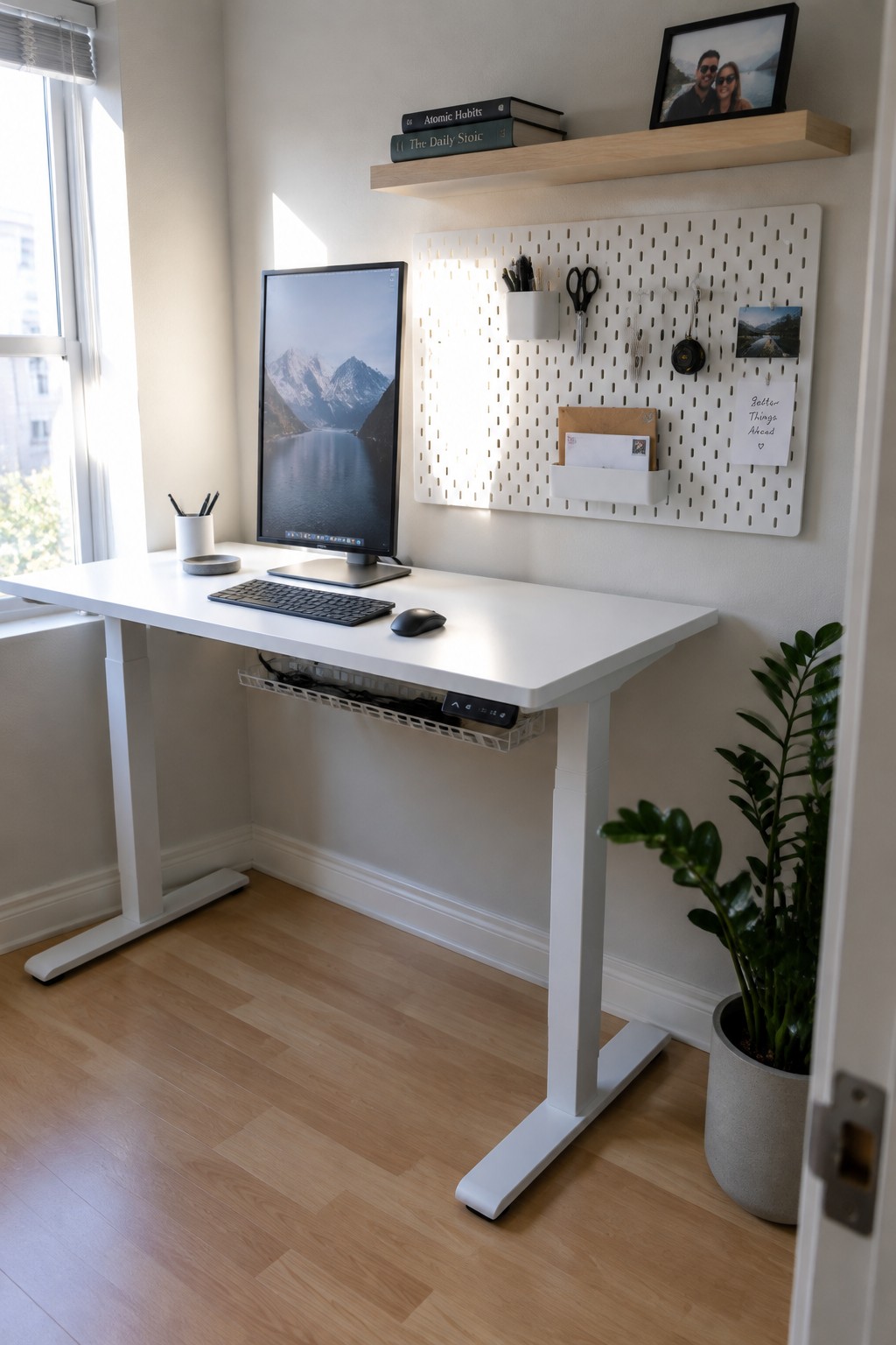 White standing desk with monitor, pegboard organizer, floating shelf, and potted plant