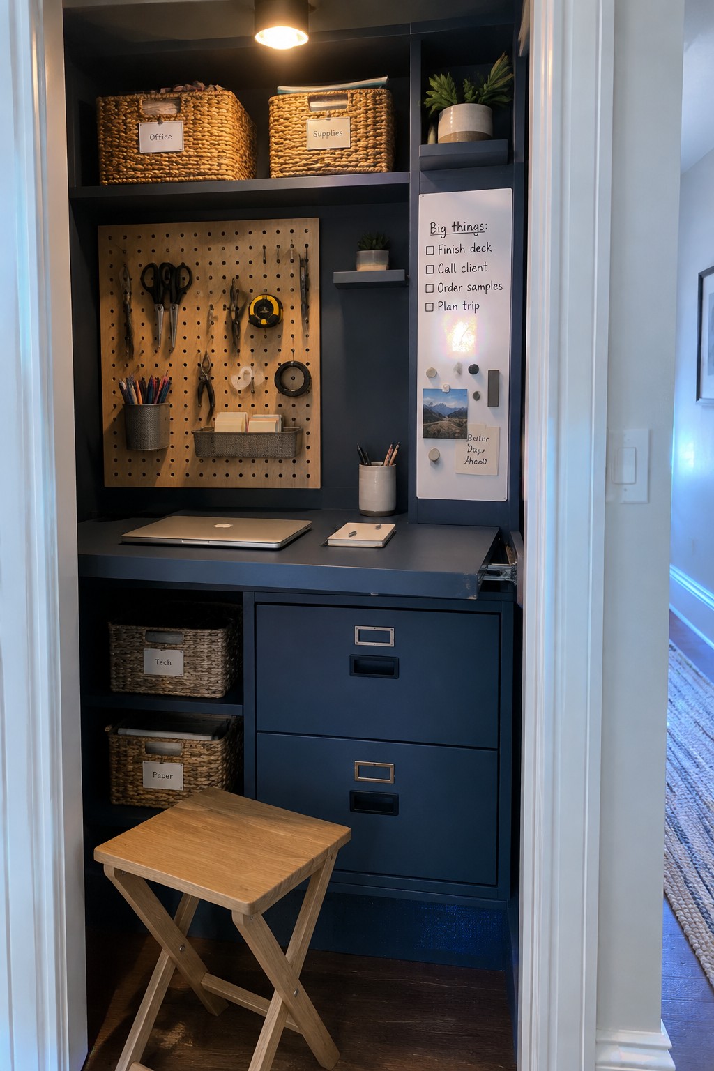 A compact built-in home office inside a closet with blue walls, a pegboard holding tools, woven storage baskets, blue filing drawers, a laptop on the desk surface, and a whiteboard with a task list.