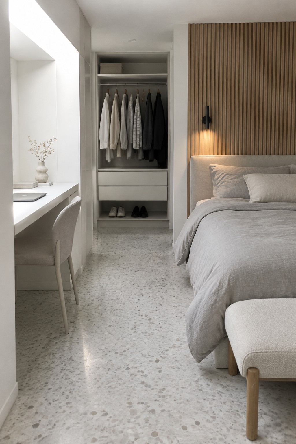 A narrow bedroom with a white desk and upholstered chair in an alcove on the left, an open closet with hanging clothes and drawers in the center, and a bed with light gray bedding on the right.