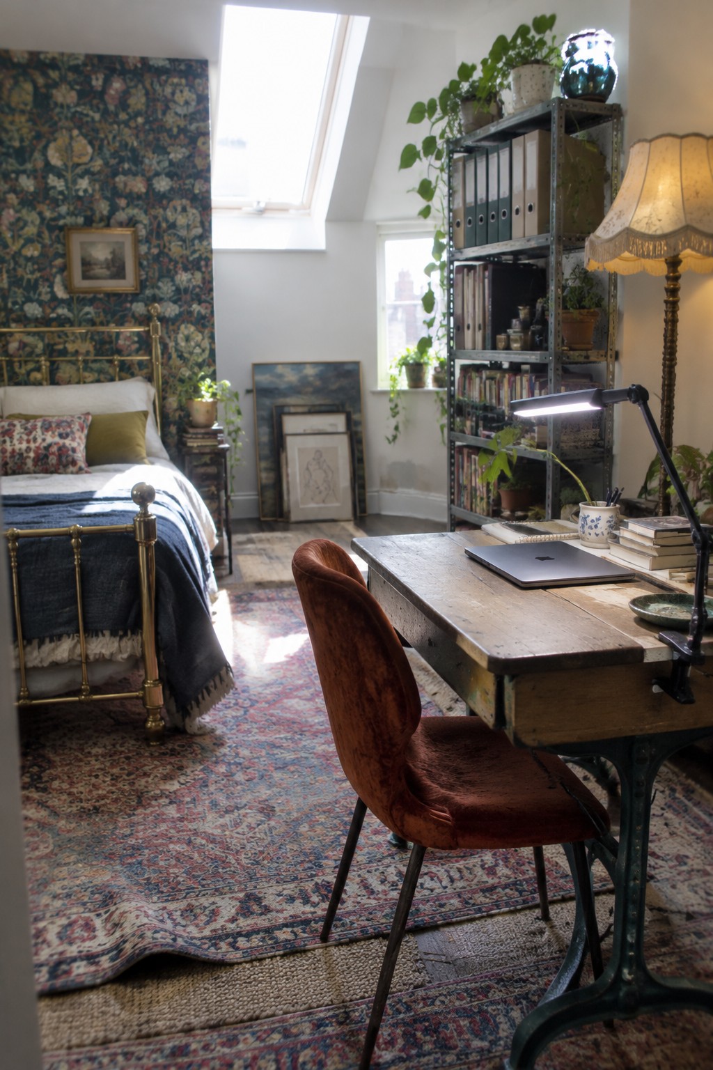 A bedroom interior showing a brass bed with patterned wallpaper on the left and a wooden desk with orange velvet chair, lit desk lamp, laptop, and metal shelving on the right under a skylight.