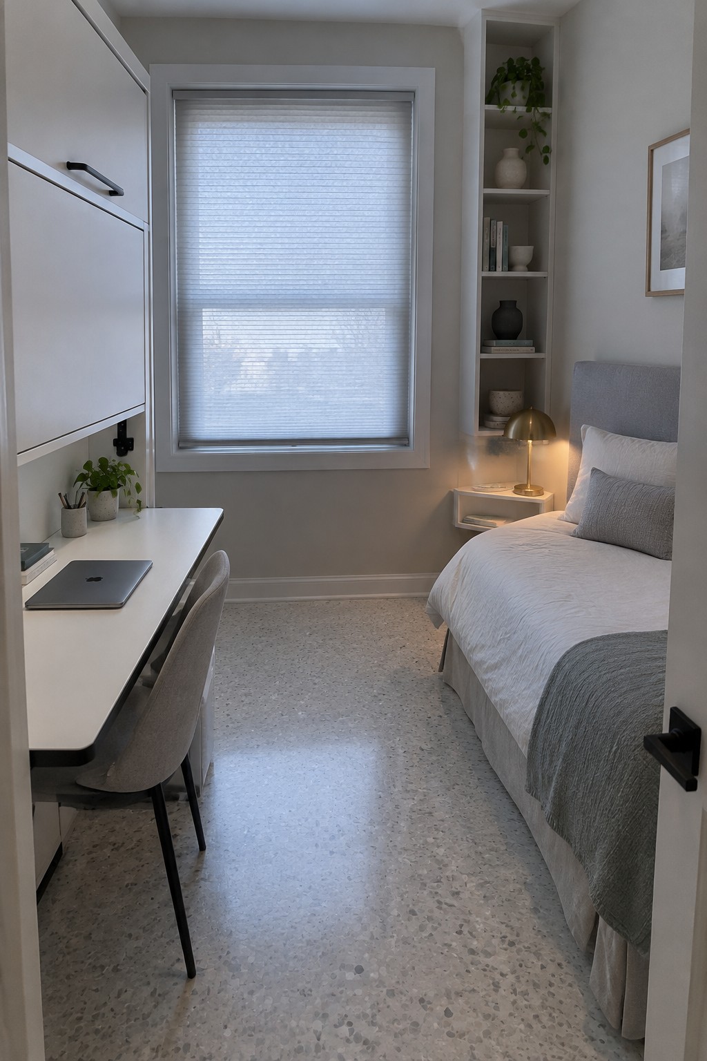 A narrow bedroom showing a white desk with a laptop and chair positioned under a window with white blinds, a bed with white and gray bedding on the right side, and built-in shelves with a lit lamp beside the bed.