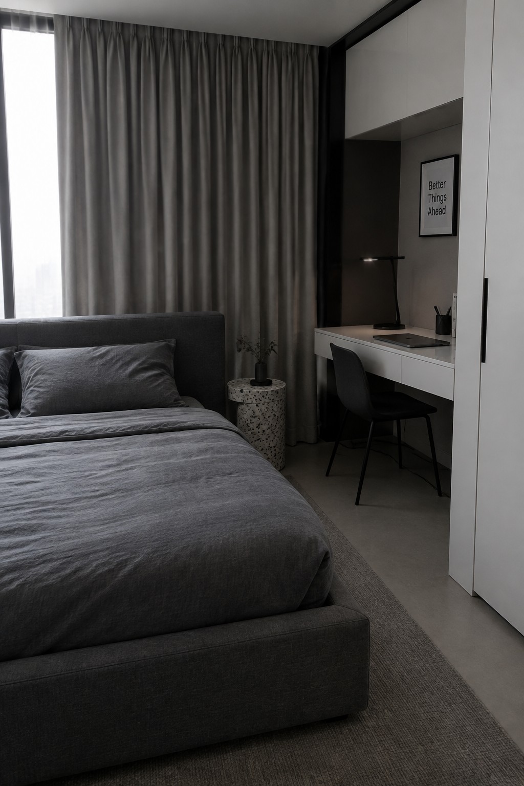 A bedroom with a large gray bed on the left and a white desk with black chair positioned beside it on the right, next to a window with gray curtains.